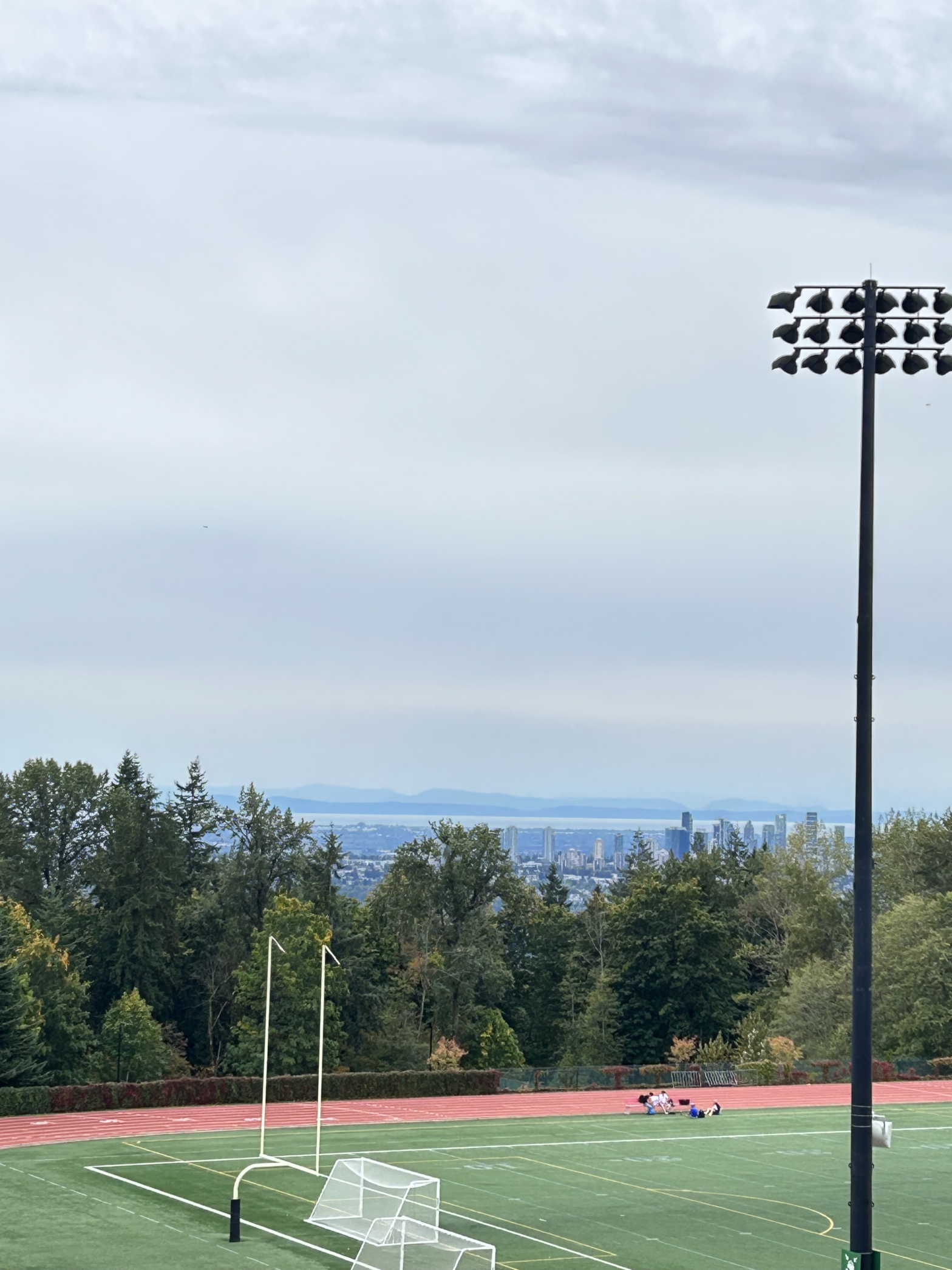 The view of a cloudy sky, trees faint mountains, the skyline, and the Terry Fox Field taken from the Rotunda Roof at Simon Fraser University (SFU) Burnaby campus.