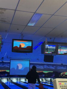 A person ready to bowl and the score tables and screens at a bowling alley.